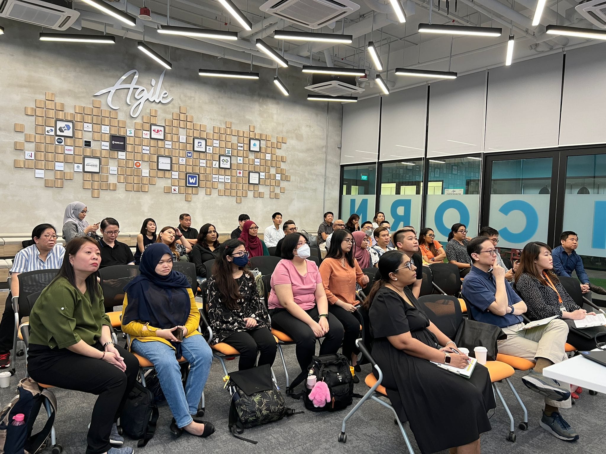A group of people seated on orange chairs in a modern room with an Agile sign and wooden block wall art.
